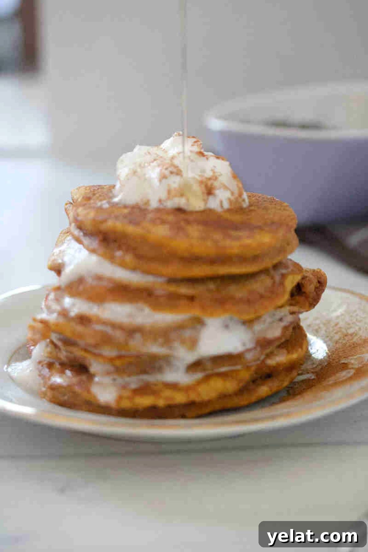 Stacked pumpkin pancakes with layers of whipped cream on a white plate and maple syrup being drizzled on top.