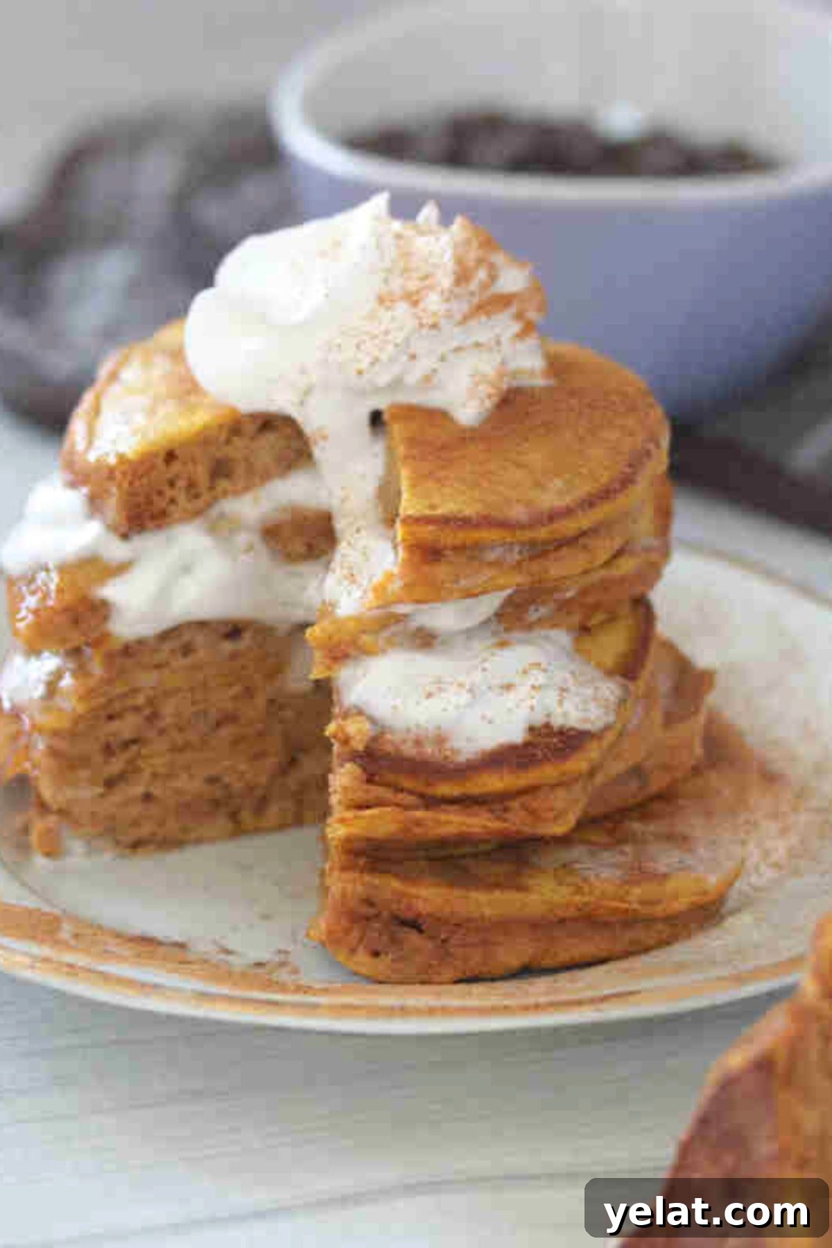 Stacked pumpkin pancakes with layers of whipped cream on a white plate with a slice out of the pancakes.