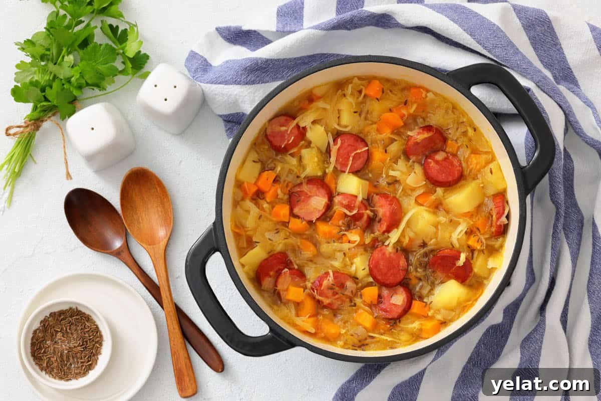 A pot of Chef Jenn’s Sauerkraut Soup with Kielbasa sits on a striped towel next to wooden spoons, bowls, fresh herbs, and salt and pepper shakers.