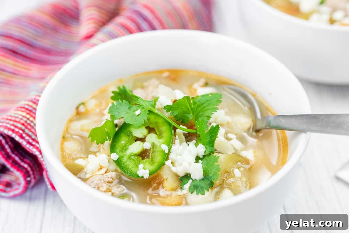 A bowl of Green pork posole soup garnished with cilantro, sliced jalapeño, and crumbled cheese, with a spoon resting inside and a striped cloth in the background.
