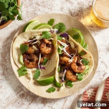 A plate of baja shrimp tacos with lime wedges, surrounded by a glass of beer, a bowl of cabbage and cilantro, and an orange and white towel.