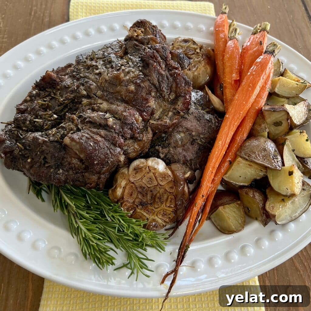 Slow-Roasted Mediterranean Lamb Shoulder Recipe 2 Slow Cooked Boneless Mediterranean Lamb Shoulder on white platter with rosemary sprigs, roasted carrots, roasted garlic and roasted potatoes.