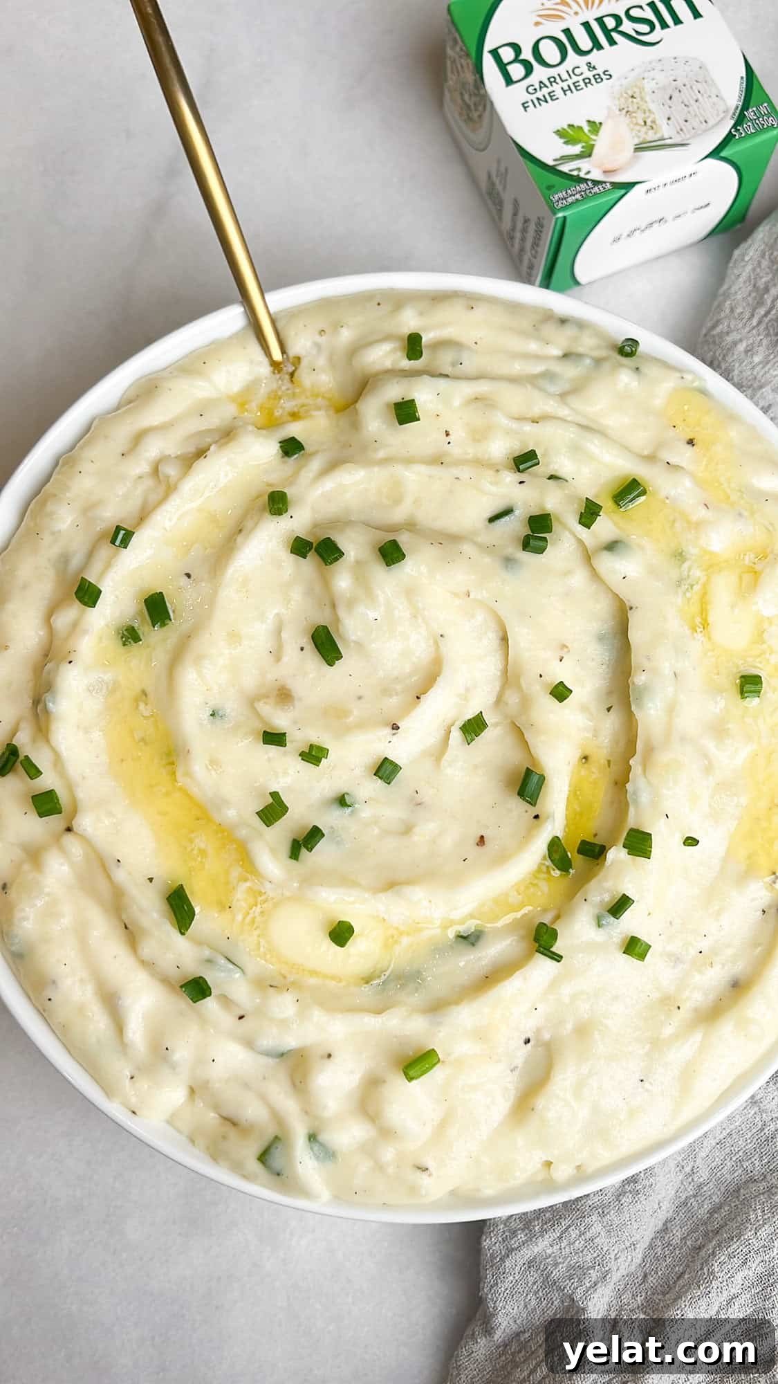 A white bowl of Boursin mashed potatoes with a gold spoon sitting beside a box of boursin cheese