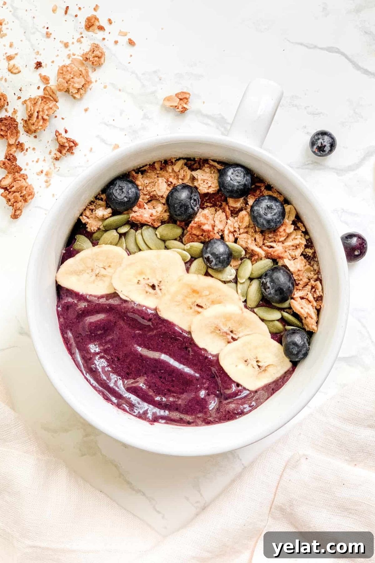 Smoothie Bowl in white bowl topped with granola, banana chips, blueberries and pepitas. Styled with gold spoon and pink linen napkin.