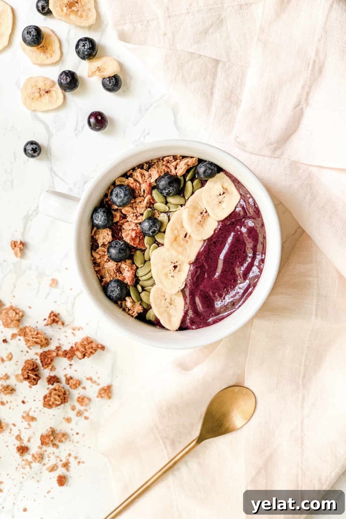Smoothie Bowl in white bowl topped with granola, banana chips, blueberries and pepitas. Styled with gold spoon and pink linen napkin.