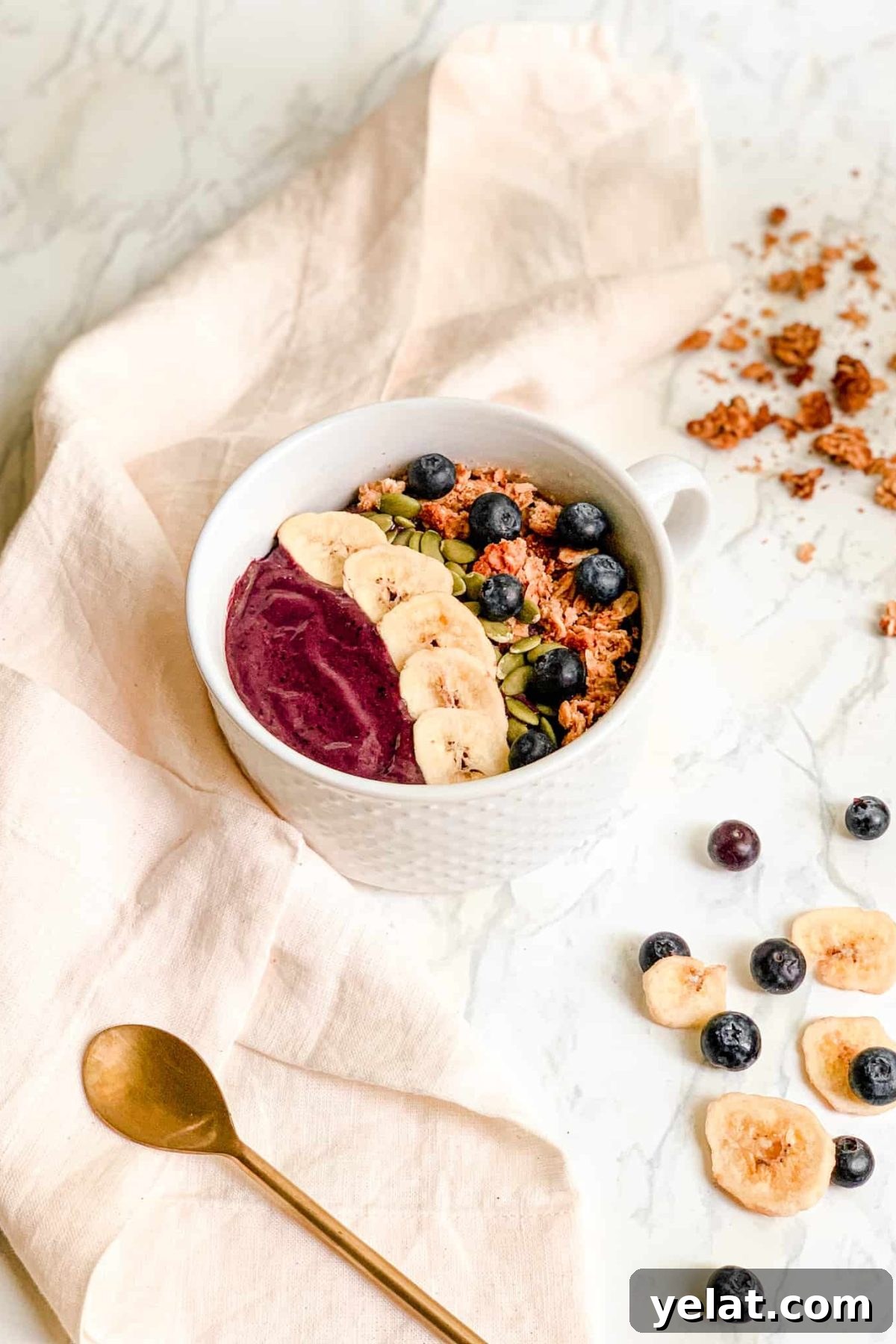 Smoothie Bowl in white bowl topped with granola, banana chips, blueberries and pepitas. Styled with gold spoon and pink linen napkin.