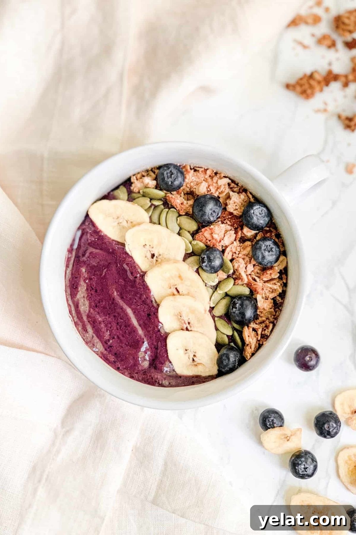 Smoothie Bowl in white bowl topped with granola, banana chips, blueberries and pepitas. Styled with gold spoon and pink linen napkin.