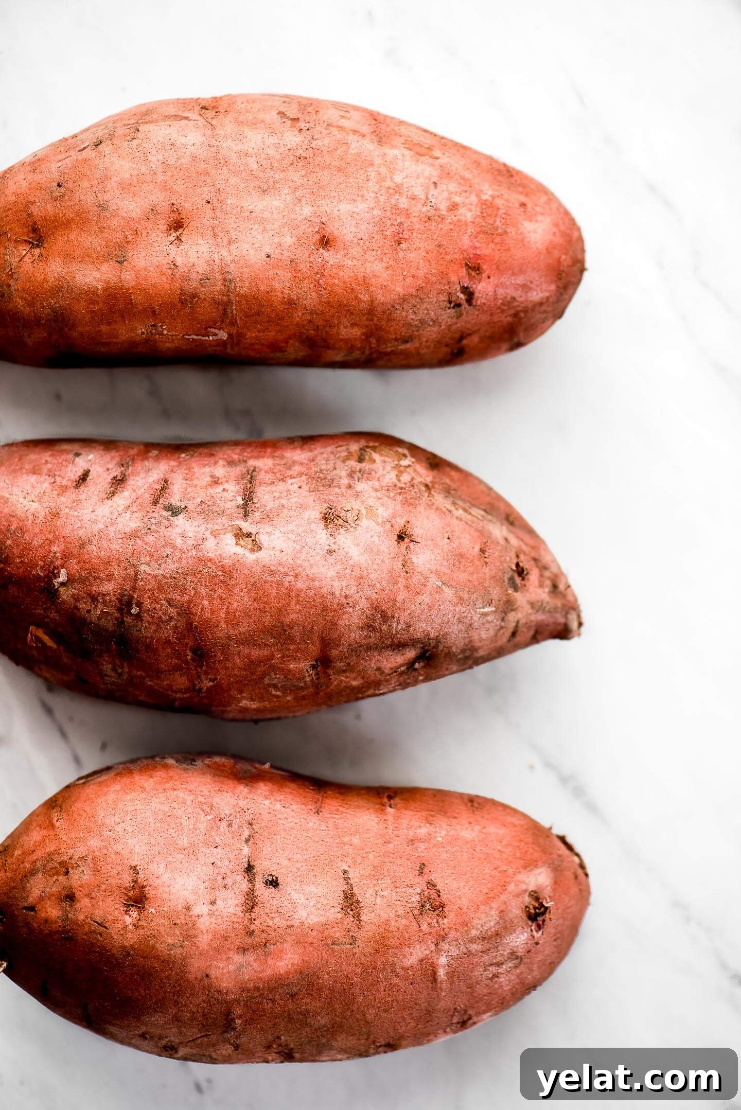Crispy Roasted Sweet Potatoes with Garlic and Herbs 3 Three raw sweet potatoes on a marble surface.