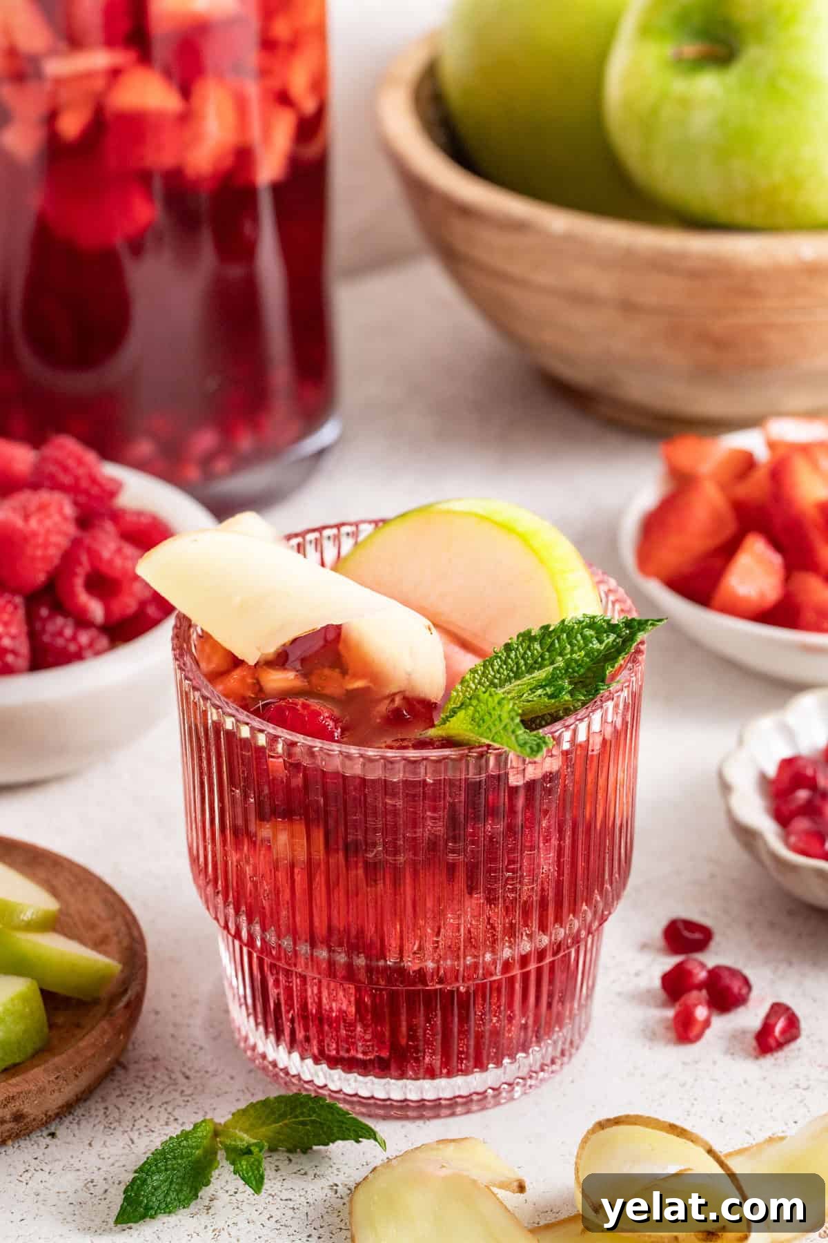 Pomegranate Virgin Sangria Recipe — Refreshing Party Drink 2 Non-alcoholic sangria in front of a pitcher with ginger, apples, and raspberries.