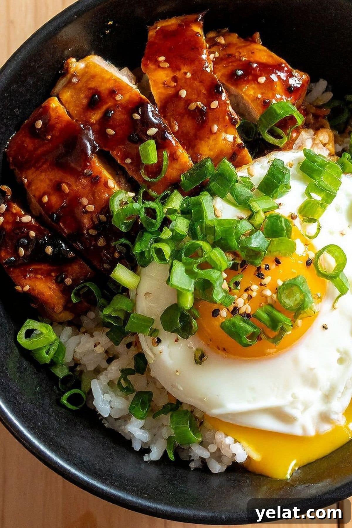 50 High-Protein Power Bowl Recipes for Every Meal 5 Teriyaki glazed chicken pieces over white rice with a fried egg, sliced green onions, and sesame seeds in a black bowl