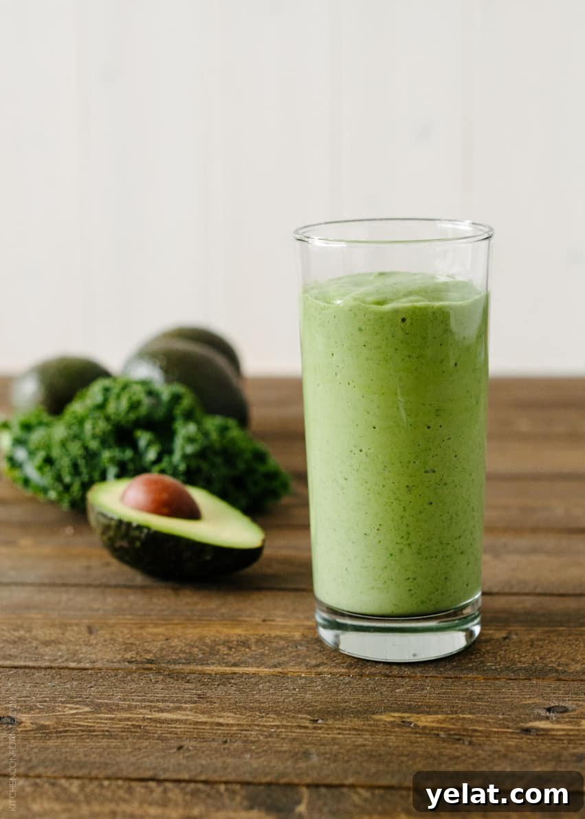 Green Avocado Kale Superfood Smoothie in a tall glass next to an avocado and kale leaves