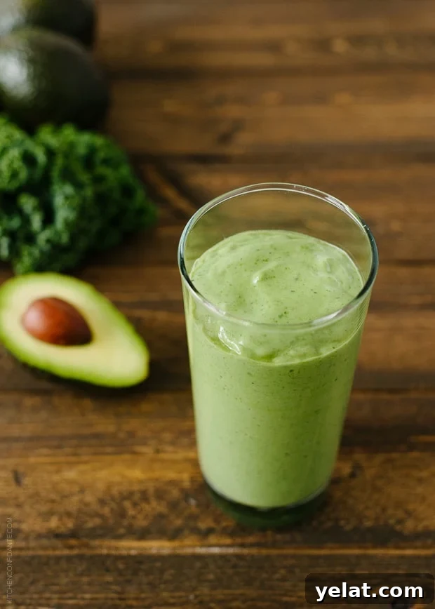 Green Avocado Kale Superfood Smoothie in a tall glass next to an avocado and kale leaves