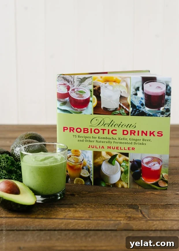 Green Avocado Kale Superfood Smoothie in a glass next to an avocado, kale leaves, and a copy of the Delicious Probiotic Drinks cookbook