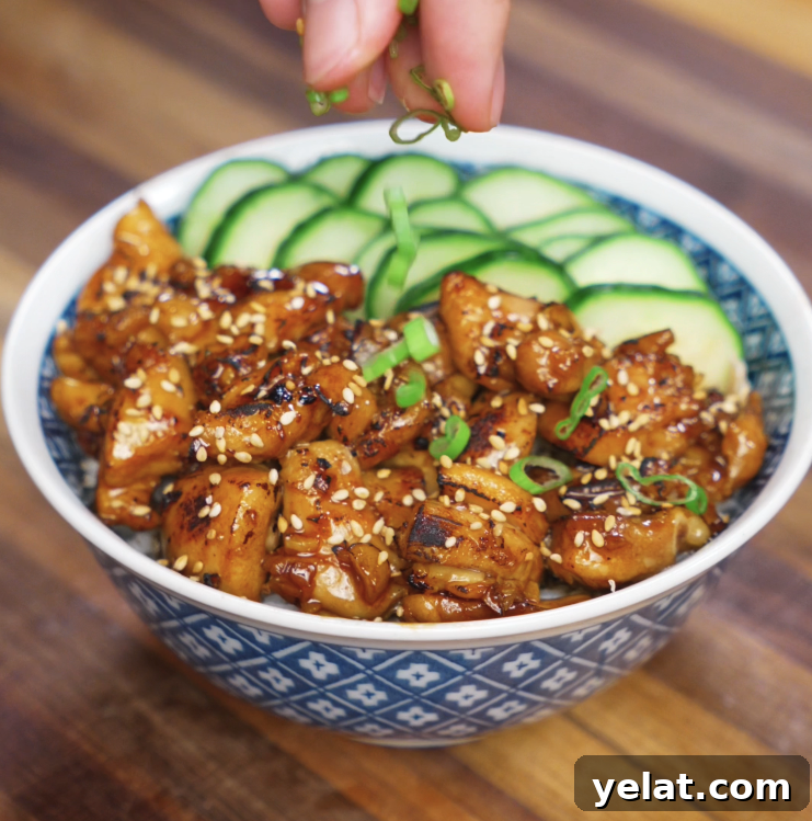 Garlic Butter Chicken Recipe with Crispy Pan-Seared Thighs 9 A hand sprinkles freshly sliced scallions over a bowl of garlic butter chicken, featuring golden, caramelized chicken glazed in a savory garlic sauce, topped with sesame seeds and served with sliced cucumber over rice.