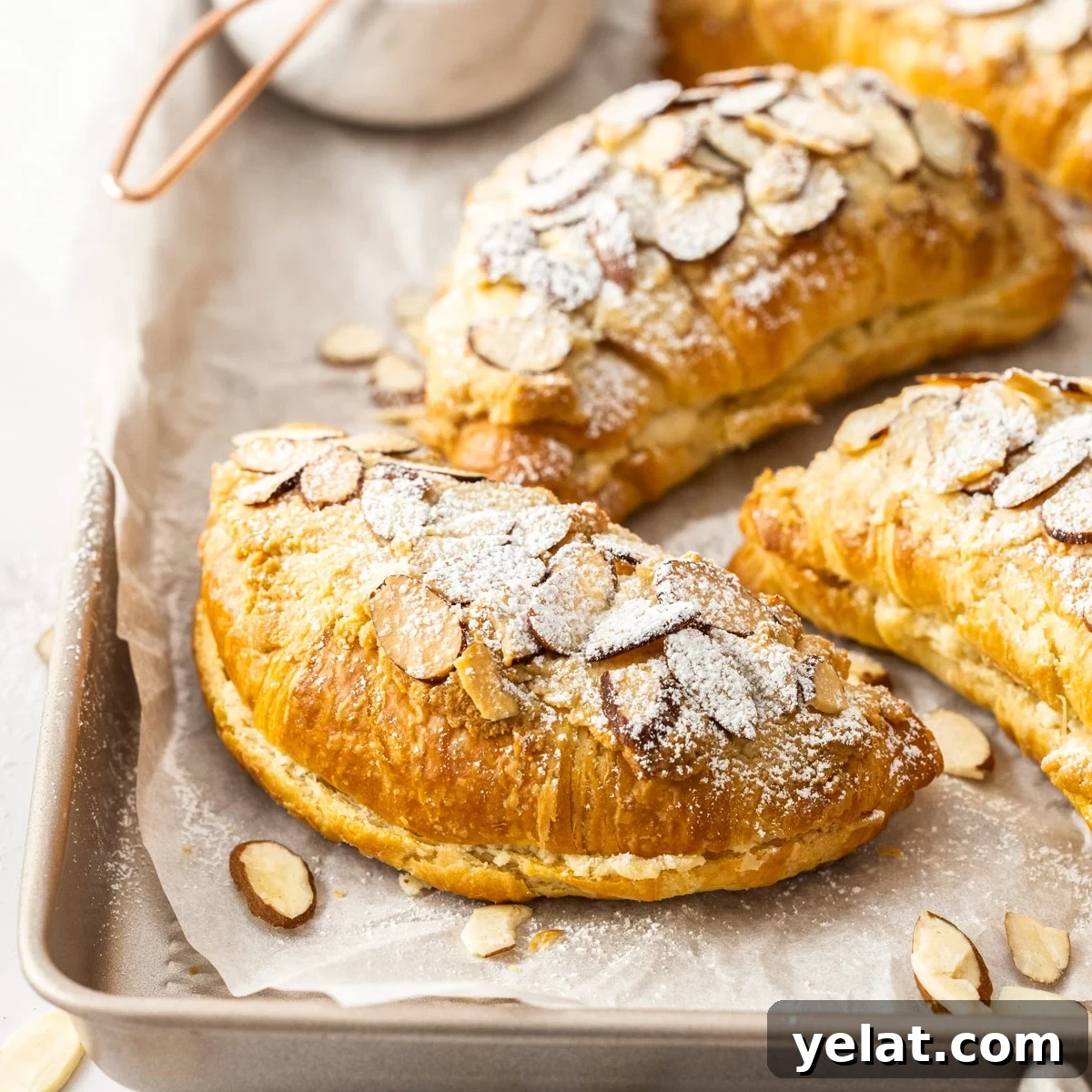 Four Almond Croissants, dusted with powdered sugar, sitting on some baking paper on a gold baking tray.
