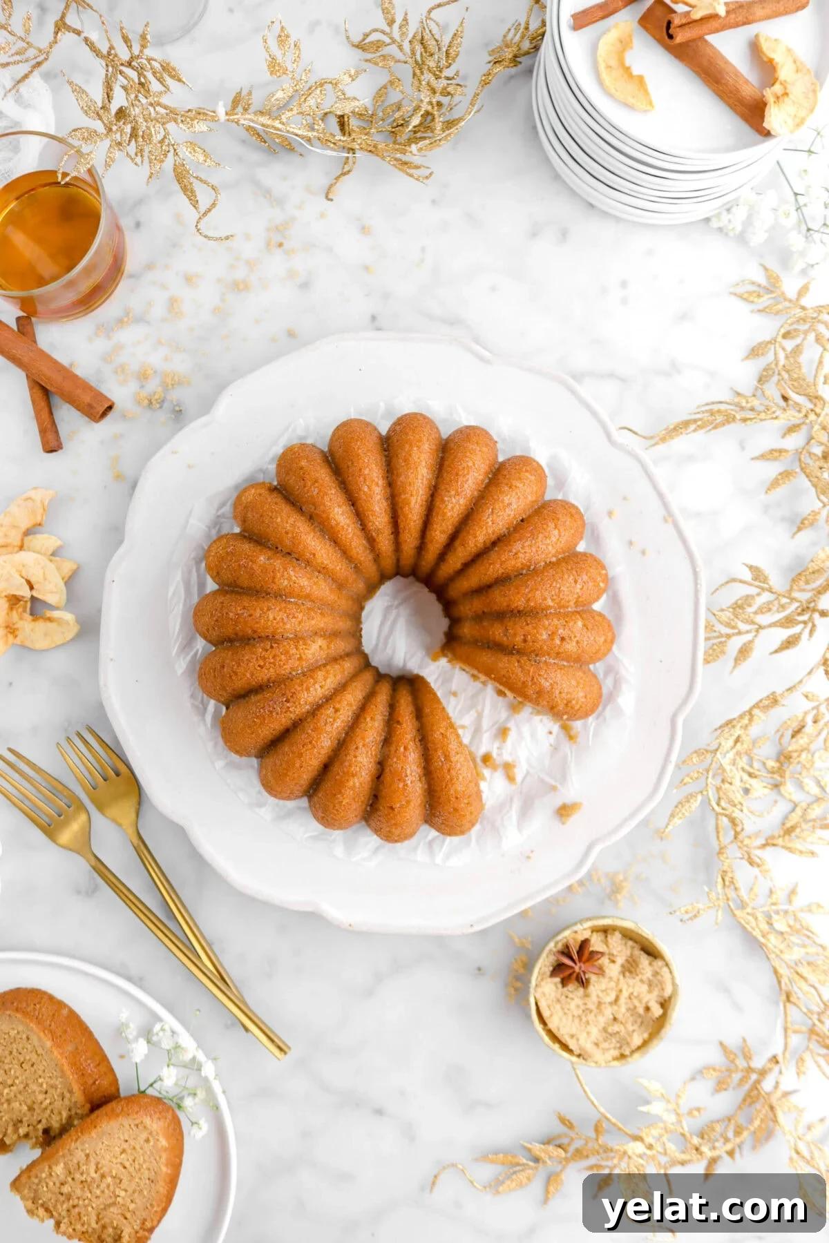 Spiced Apple Cider Bourbon Bundt Cake Recipe for Fall Desserts 5 overhead shot of bundt cake with gold leaf garland around, two slices on white plate with white flowers, gold bowl of brown sugar and two forks beside on marble surface