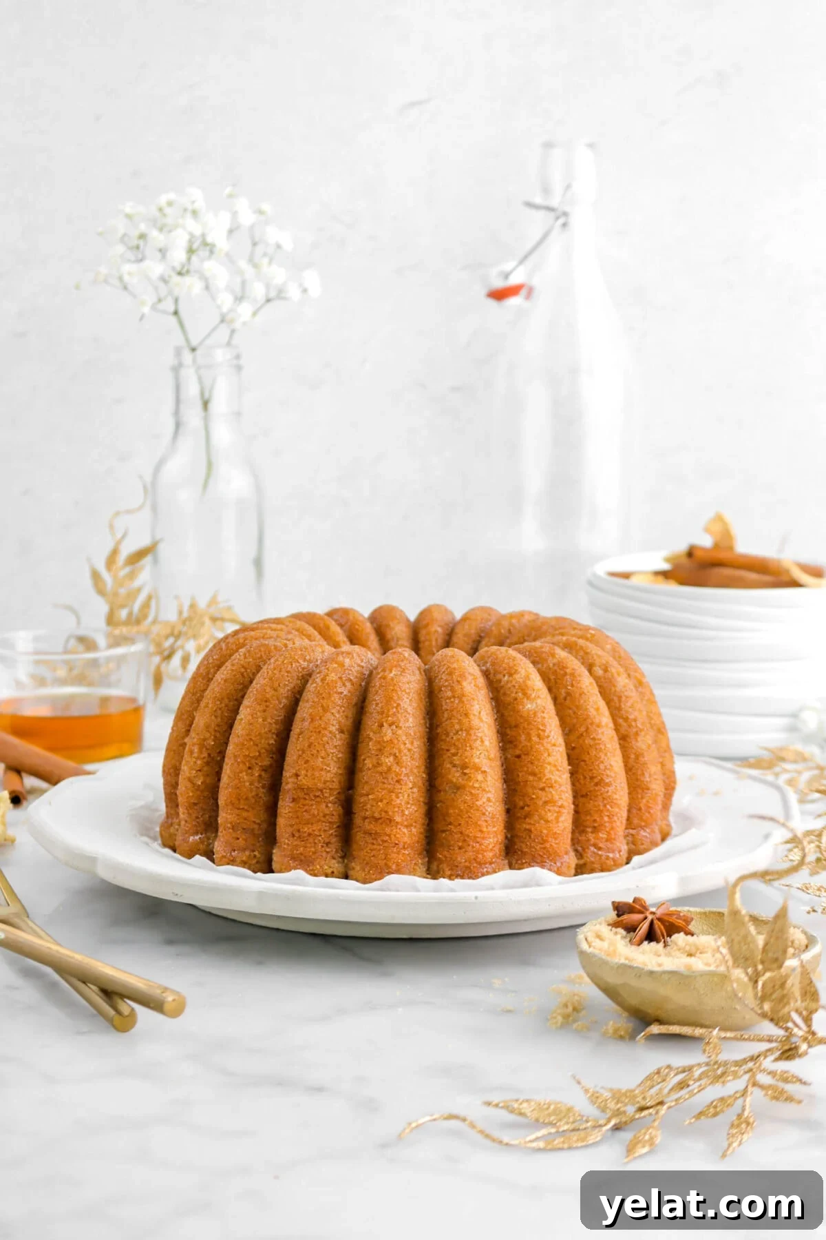 Spiced Apple Cider Bourbon Bundt Cake Recipe for Fall Desserts 6 front shot of bundt cake on grey plate with gold garlic, glass of white flowers behind, and stack of plates.