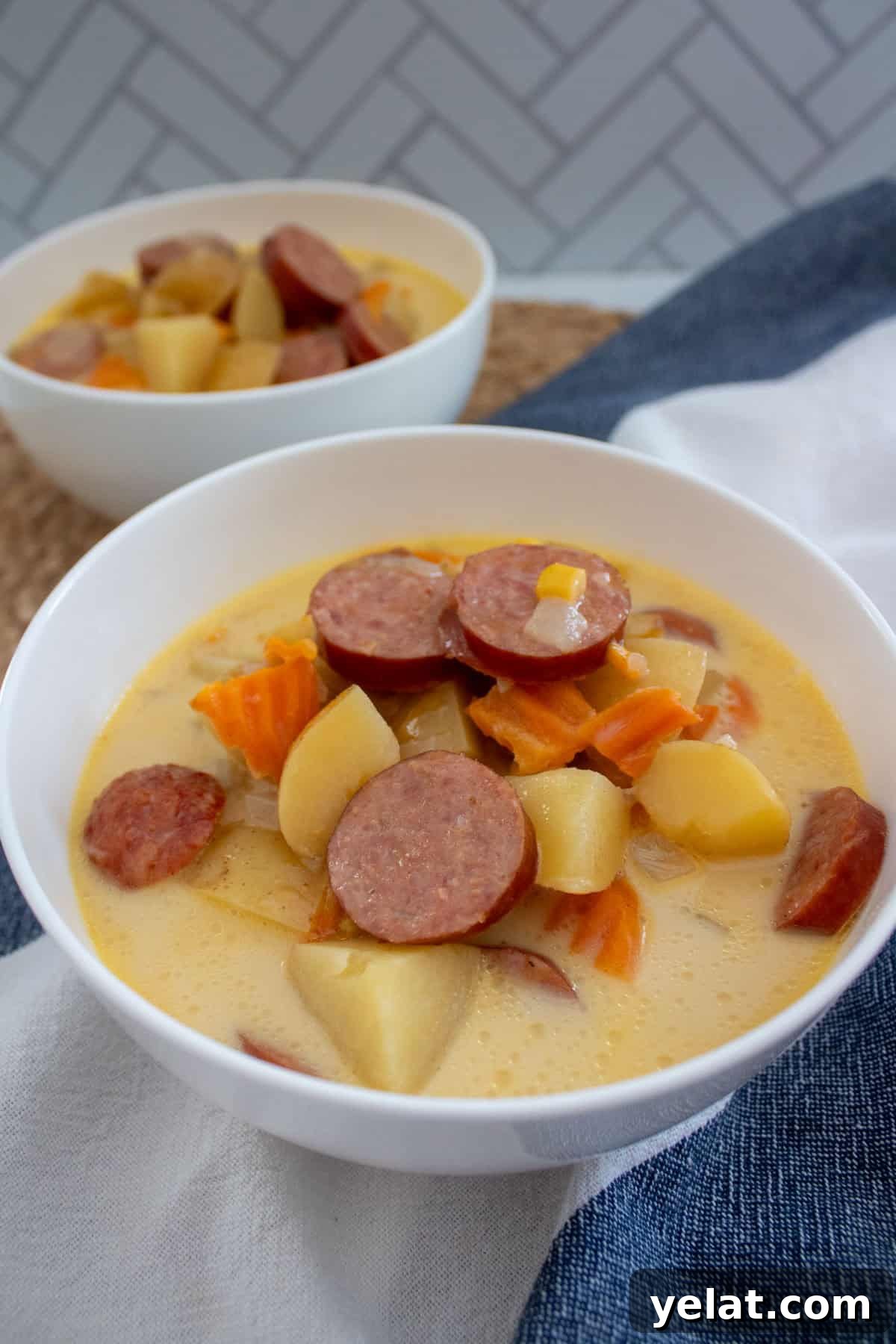 A white bowl filled with creamy soup containing sliced sausage, potatoes, carrots, and corn, with another similar bowl in the background. The scene includes a white napkin and a woven placemat.