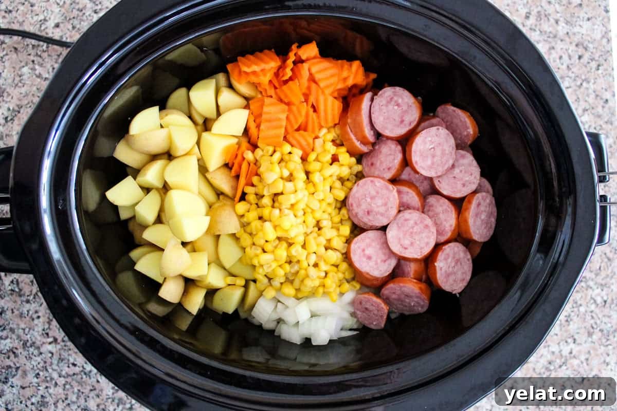 Chopped potatoes, carrots, corn, onions, and sliced sausage are arranged in sections inside a black slow cooker on a speckled countertop.
