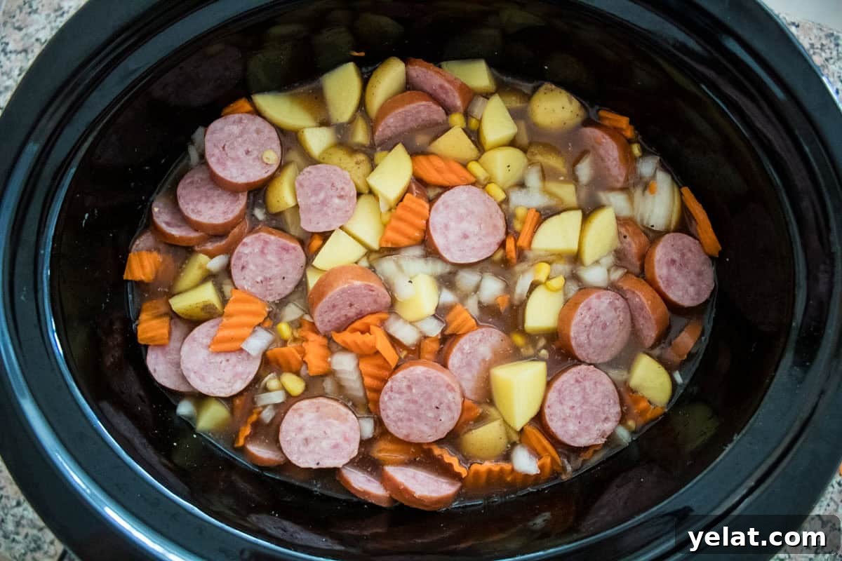 Sliced sausage, potatoes, carrots, onions, and broth in a black slow cooker, ready to be cooked into a soup.