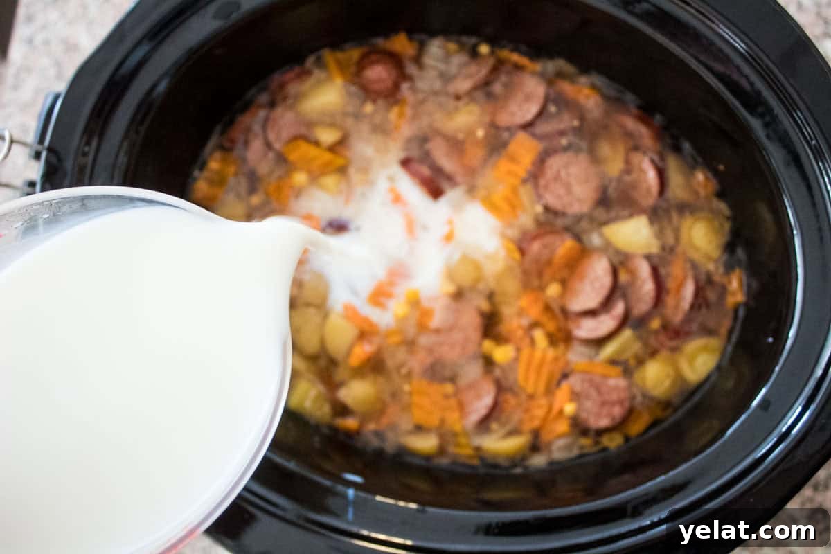 Milk is being poured from a measuring cup into a slow cooker filled with sliced sausage, carrots, potatoes, and broth.