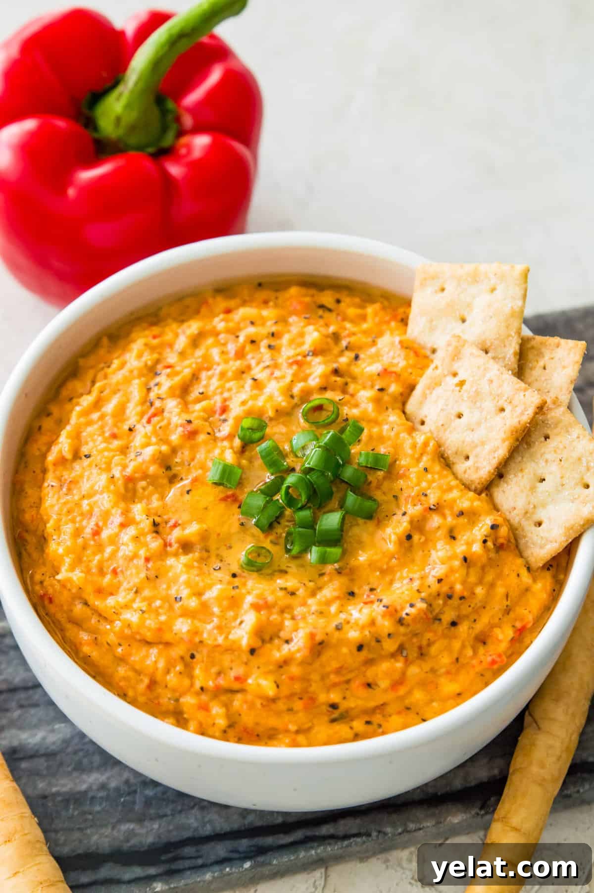 Roasted Red Pepper Cashew-Parsnip Dip (Vegan) 2 A bowl of roasted red pepper dip garnished with chopped green onion with four crackers in it.