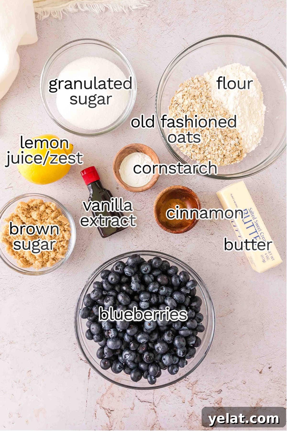 Bowls containing ingredients for Blueberry Crumble Pie—granulated sugar, flour, old fashioned oats, brown sugar, cornstarch, cinnamon, butter, vanilla extract, lemon juice/zest, and blueberries—are arranged on a countertop.