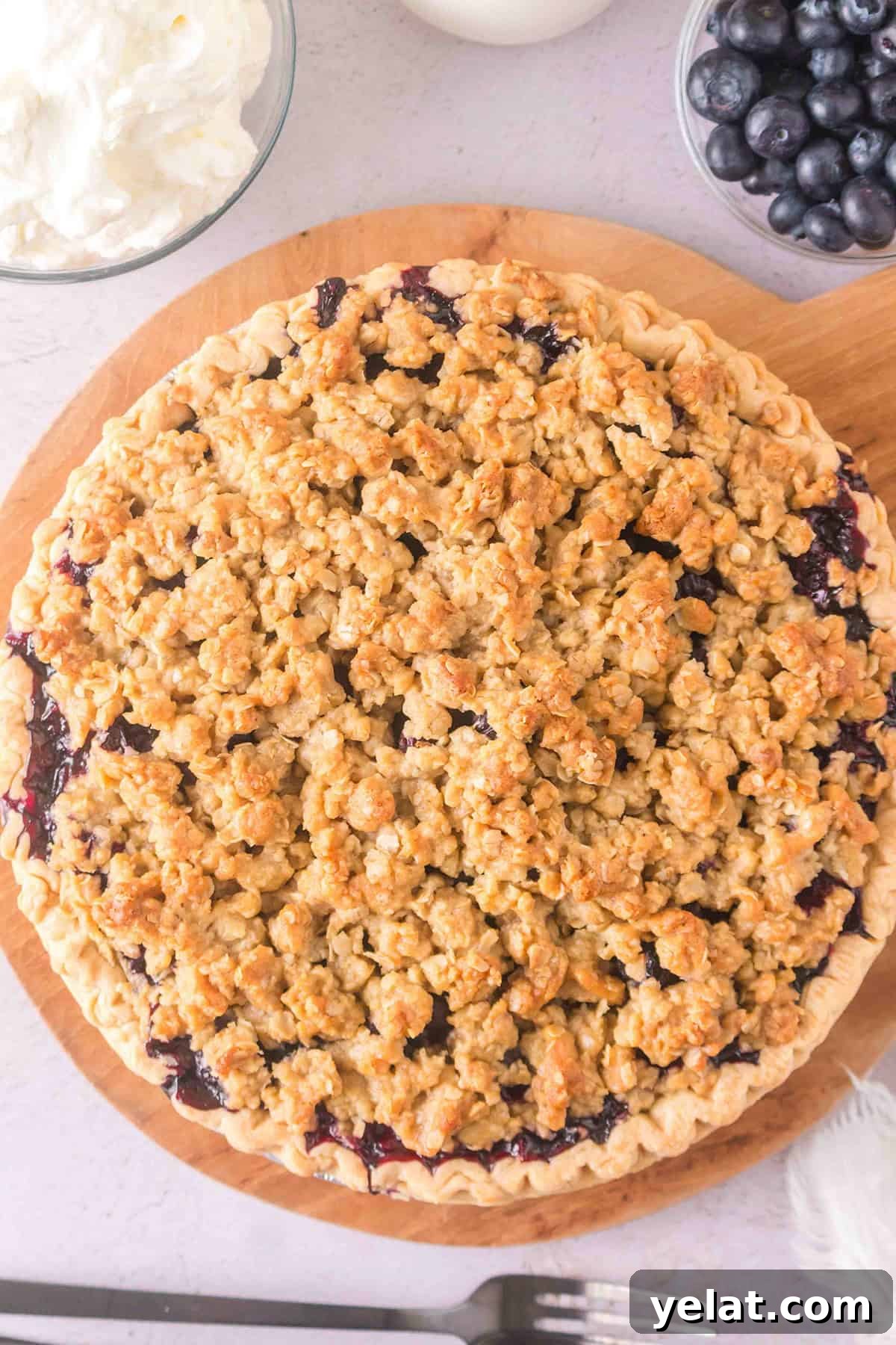 A baked blueberry pie with a golden crumble topping sits on a wooden board, accompanied by a bowl of whipped cream and fresh blueberries nearby.