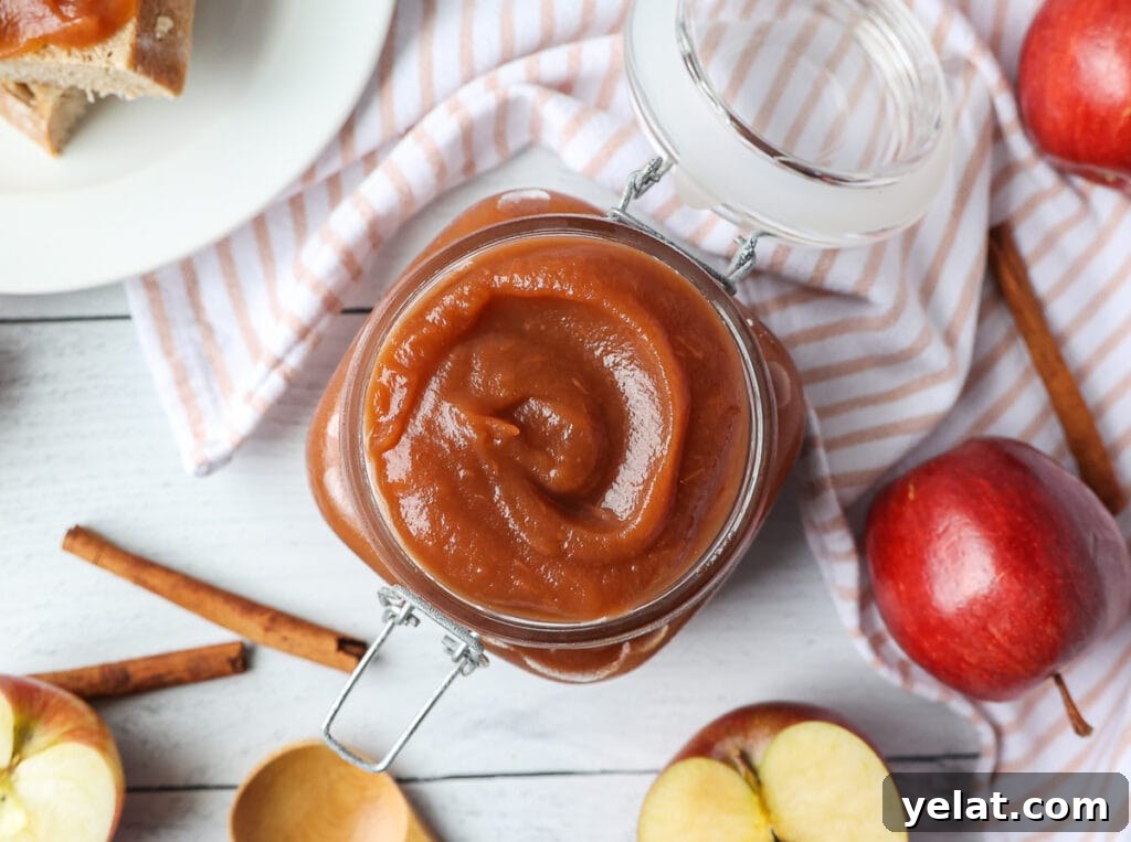 A top view of a jar of apple butter. Cinnamon sticks and apples surrounding the jar with a napkin.