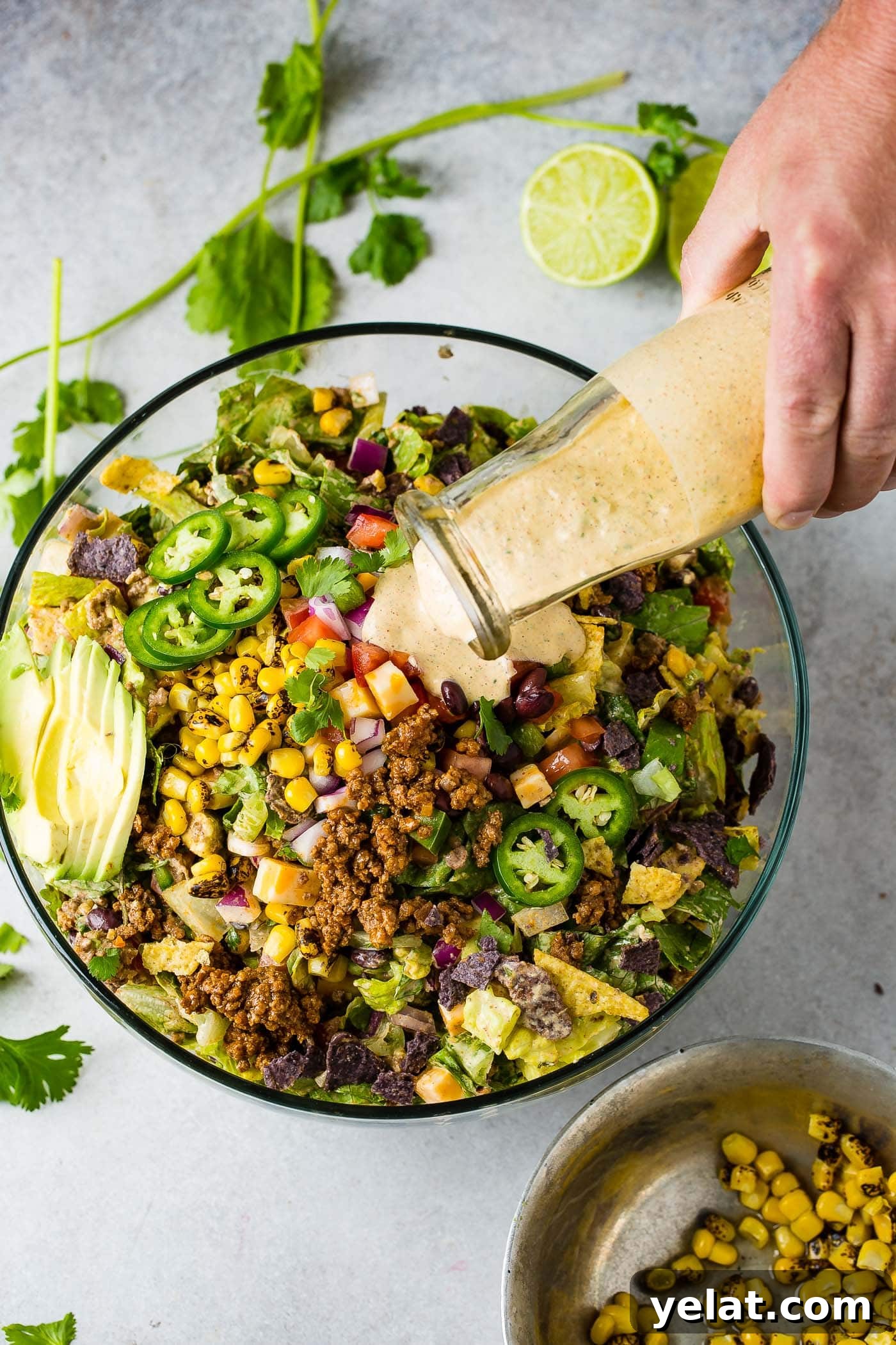 Tex-Mex Chopped Ground Beef Taco Salad Recipe 10 A glass bowl filled with tex‑mex taco salad. The salad has avocado slices, corn, taco meat, tomatillos, tomatoes, and chips. Dressing is being poured from a glass container onto the salad.