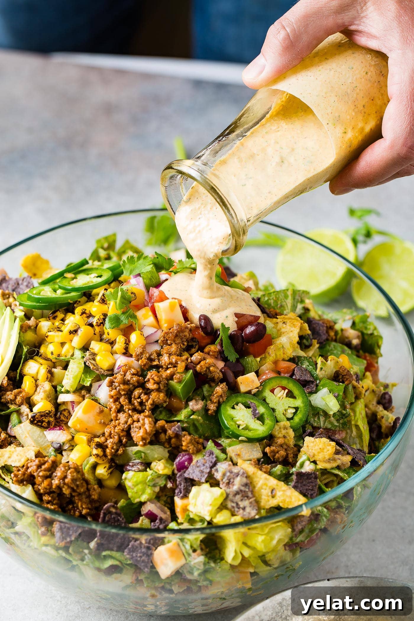 Tex-Mex Chopped Ground Beef Taco Salad Recipe 6 A glass bowl filled with tex‑mex taco salad. A dressing is being poured over the salad. The salad is packed with corn, tomatillos, tomatoes, lettuce taco meat, and a sliced avocado is on the edge of the salad.