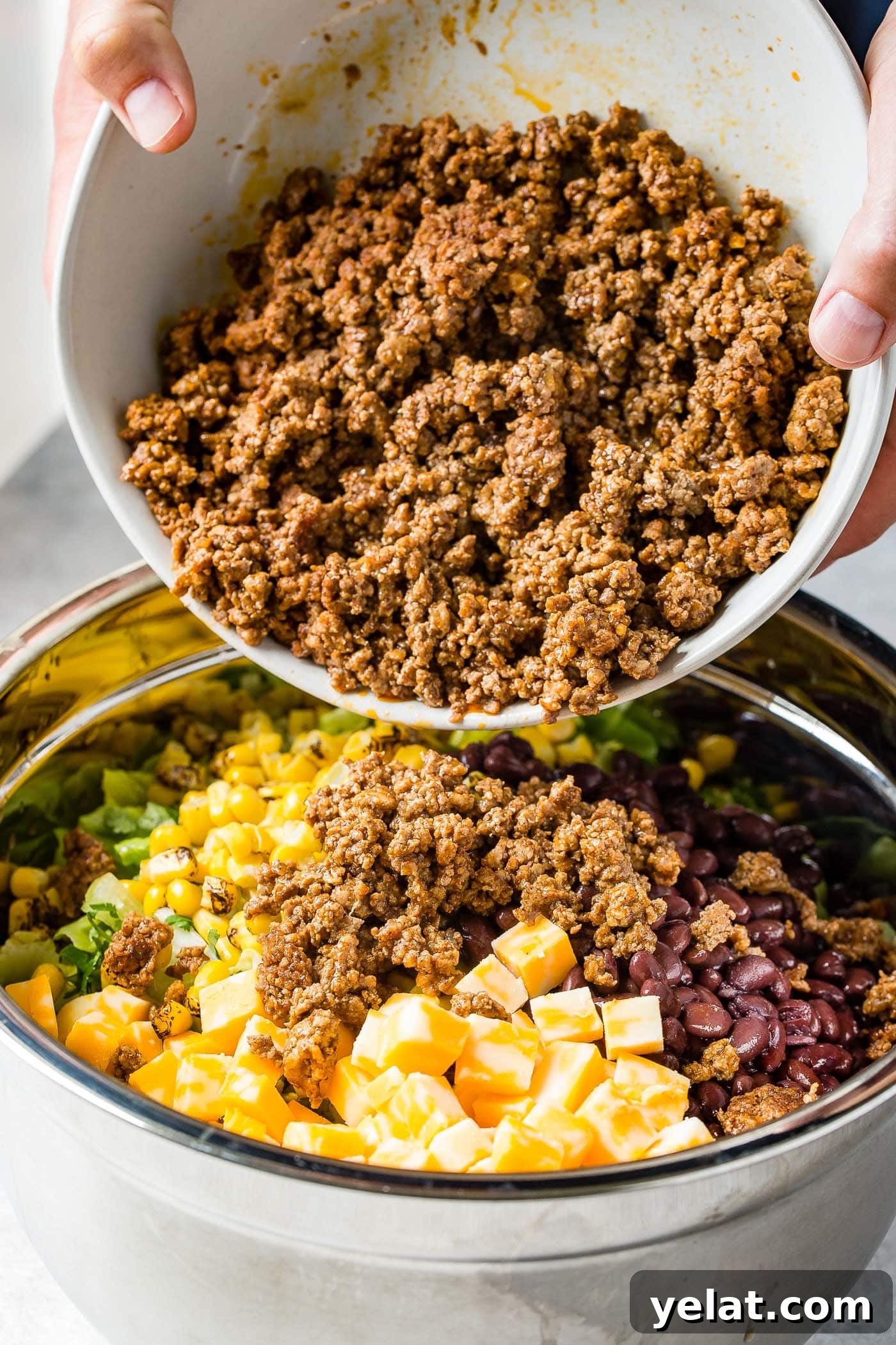 Tex-Mex Chopped Ground Beef Taco Salad Recipe 8 taco meat being poured into a bowl with corn, tomatillos, cheese cubes and chopped onions.