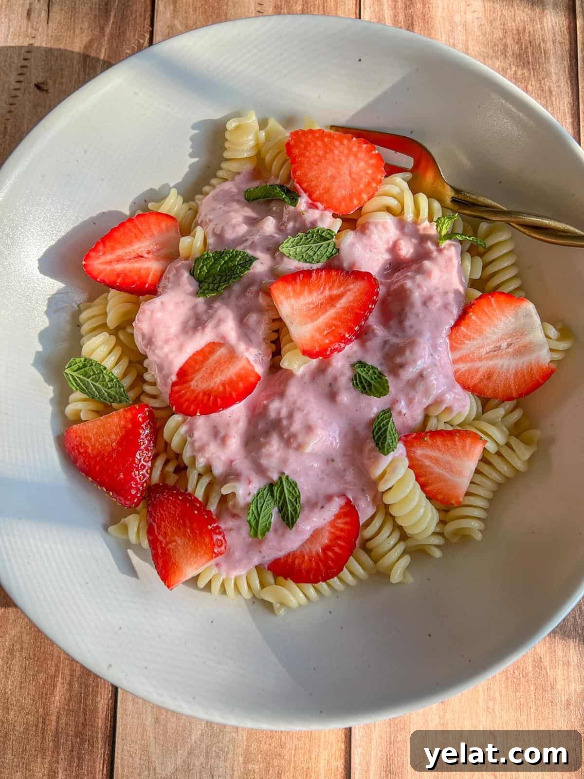 Polish Strawberry Pasta Recipe: Sweet Summer Noodles with Strawberries 2 A white bowl with spiral pasta topped with pink strawberry cream sauce, fresh strawberry slices, and mint leaves, set on a wooden surface with a gold fork.