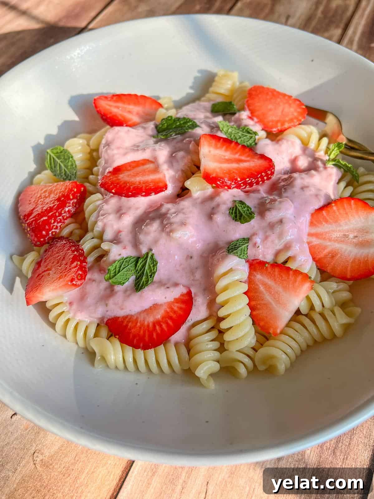Polish Strawberry Pasta Recipe: Sweet Summer Noodles with Strawberries 3 A white plate of fusilli pasta topped with pink strawberry sauce, fresh strawberry slices, and mint leaves, set on a wooden surface in natural light.