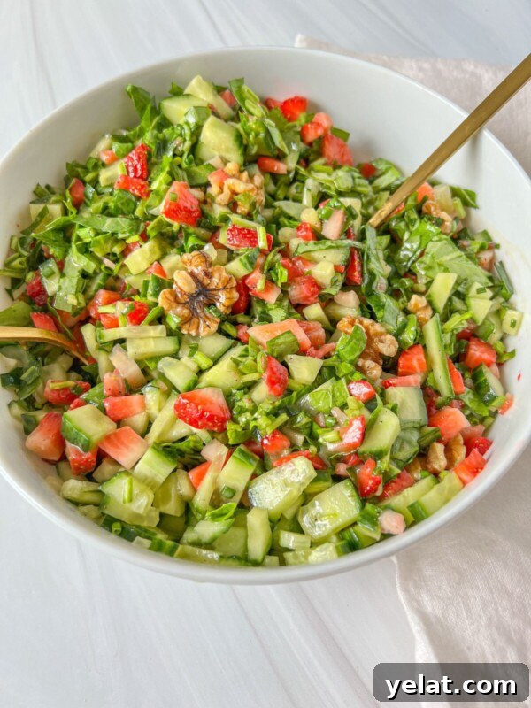 Polish Strawberry Pasta Recipe: Sweet Summer Noodles with Strawberries 4 A white bowl filled with a fresh Jennifer Aniston Salad made of chopped cucumbers, strawberries, greens, and walnuts, with a gold spoon and napkin set on a light surface.