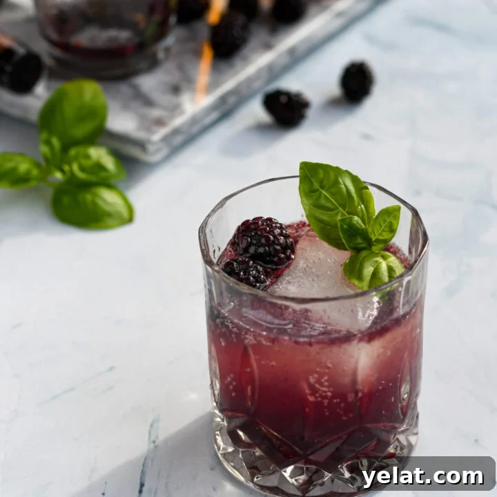 an old fashioned glass filled with a purple blackberry bourbon smash garnished with a large ice cube, blackberries, and basil