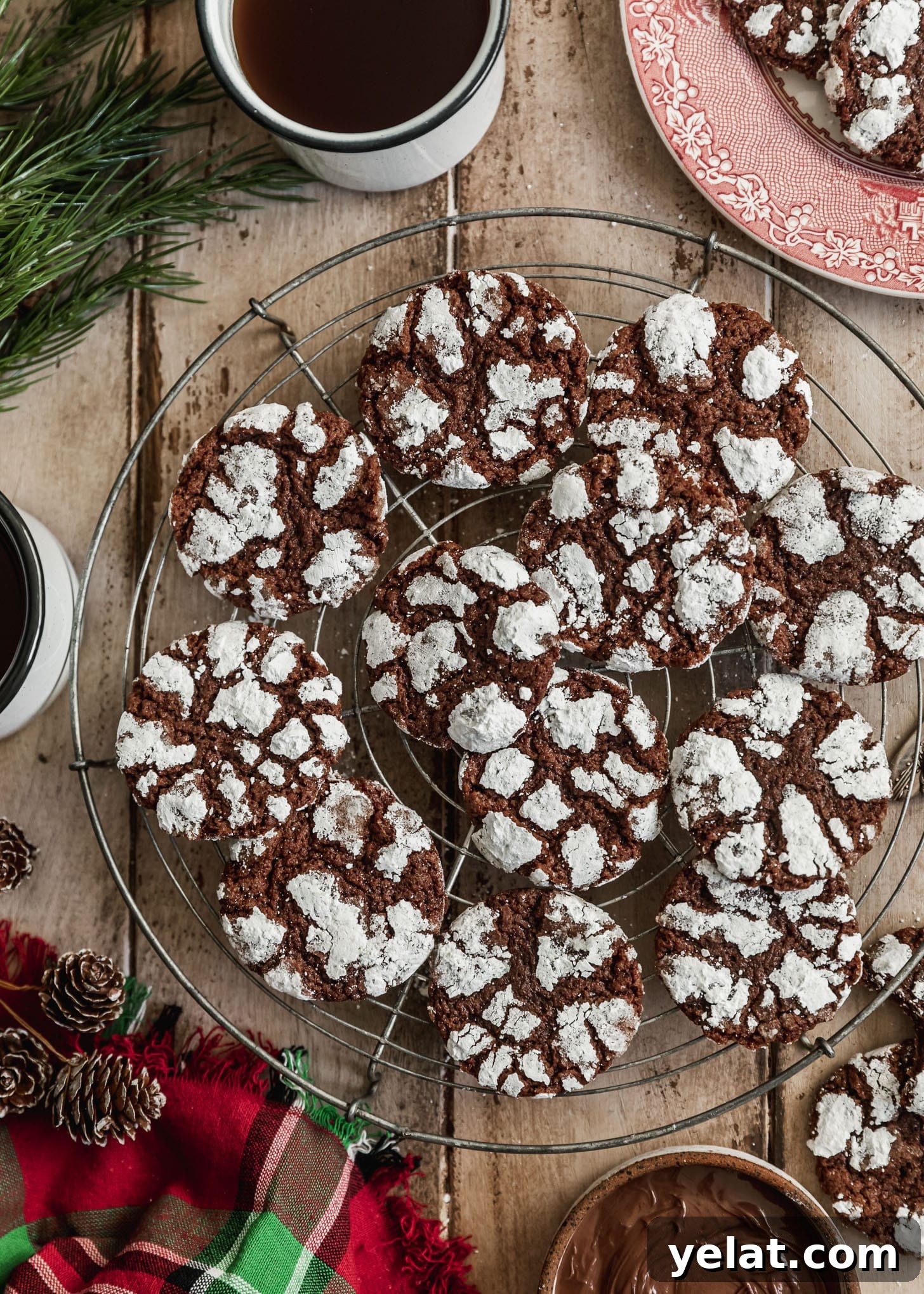 Fudgy Mocha Crinkle Cookies Recipe for Rich Chocolate Coffee Cookies 8 A wire rack of mocha crinkle cookies on a wood table next to a red plaid linen, pinecones, a white bowl of Nutella, and white mugs of coffee.