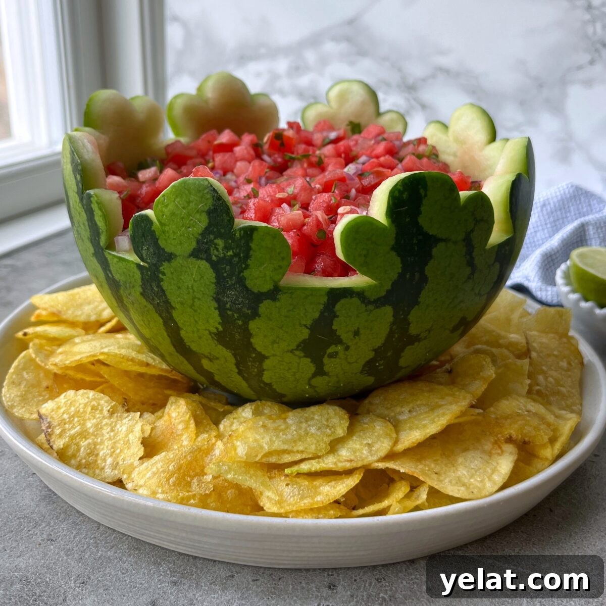 Fresh Watermelon Salsa Recipe for Summer Parties 3 A carved watermelon bowl filled with Watermelon Salsa sits on a plate surrounded by potato chips on a gray countertop.