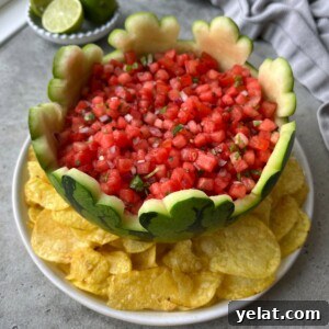 Fresh Watermelon Salsa Recipe for Summer Parties 8 A hollowed-out watermelon bowl filled with fresh watermelon salsa sits on a white plate surrounded by yellow tortilla chips. Lime wedges and a gray cloth are nearby.